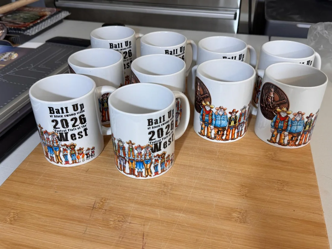 White ceramic event mugs with matching cowboy illustrations and event lettering on a workbench